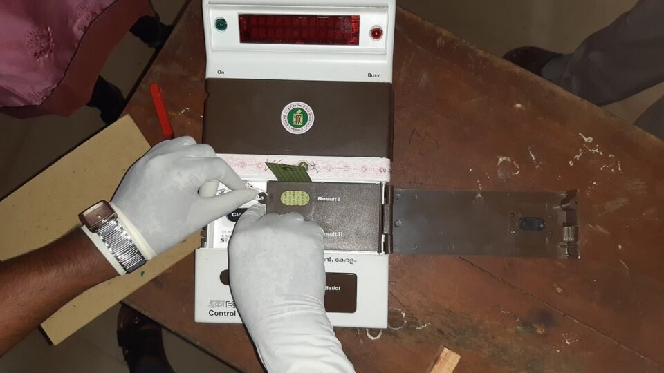 दस्ताने पहने हाथों में इलेक्ट्रॉनिक वोटिंग मशीन (EVM) की कंट्रोल यूनिट