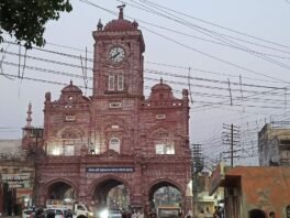 मेरठ शहर का लाल रंग का घंटाघर और सामने सड़क पर चलते वाहन